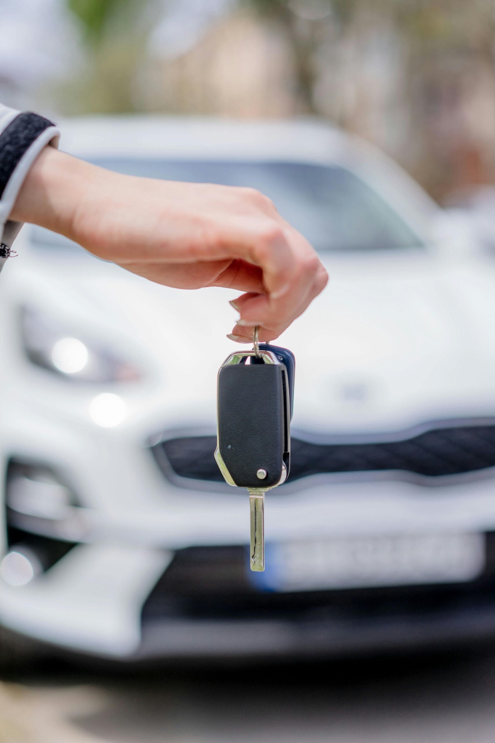 Close-up of a hand holding a car key, with a white car in the background outdoors.